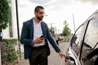 © kleberpicui - Urban professional using smartphone to request or access ride hailing transportation beside a parked car in a modern city environment, representing app mobility and daily commuting.