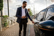 © kleberpicui - Urban professional using smartphone to request or access ride hailing transportation beside a parked car in a modern city environment, representing app mobility and daily commuting.
