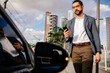 © kleberpicui - Urban professional using smartphone to request or access ride hailing transportation beside a parked car in a modern city environment, representing app mobility and daily commuting.