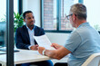© Mediaphotos - Adult man handing document to young Black man during business meeting in office, both sitting at desk, computer monitor in background