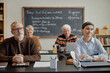 © Seventyfour - Group of senior Caucasian men and women attending class, sitting at desks and listening attentively, blackboard with handwritten notes in background, education setting