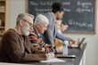 © Seventyfour - Group of middle aged and senior Caucasian men sitting at desk writing in notebooks, while Black woman standing nearby assisting with laptop during adult education class