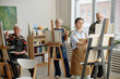 © Seventyfour - Group of senior Caucasian men and women participating in painting class, standing at easels and focusing on creating artwork in educational setting with natural light from windows