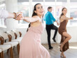 © JackF - Cheerful young brunette in light pink flowing dress practicing swing dance steps with male partner in sunny choreographic studio. Social dancing concept