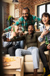 © DC Studio - Room full of excitement as black woman celebrates video game win surrounded by diverse friends. Cheerful male and female individuals enjoy weekend gaming hangout, looking at TV with joy at home.