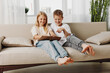 © SHOTPRIME STUDIO - Two happy children sitting on a couch using a tablet together in a bright living room, enjoying digital entertainment and shared moments.