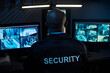 © pressmaster - Black man middle aged sitting at security desk monitoring multiple surveillance camera feeds on computer screens in control room, back of head and uniform with security label visible