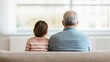 © Jack - A photorealistic contemporary living room scene illustrating connection bridging generations, showing a grandparent and child sitting side by side on a neutral fabric sofa viewed from behind.