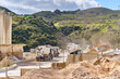 © Travel 'n' Lifestyle - Manilva, Spain - 18 February 2026: View of a concrete plant with trucks, industrial buildings, and earthy tones contrasting with vibrant green hillsides, under a bright, expansive sky.