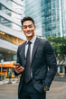 © Kowit - A confident businessman in a suit smiles while holding a smartphone in an urban city environment with modern glass buildings in the background.