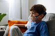 © kleberpicui - Young child receiving nebulizer treatment at home while sitting on sofa, representing pediatric respiratory care, asthma management and family health routine.