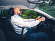 © mark_gusev - Bald man with grey beard is a car driver. Male in light blue shirt holding driving wheel firmly, green forest background.