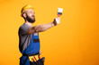 © DC Studio - Male contractor in hardhat extends paintbrush forward, appearing to inspect coverage or gesture for paint direction. White repairman stands representing painting work or home renovation services.