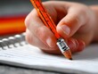 © Samorn - A childs hand grips an orange pencil writing on lined paper in a spiral notebook, getting ready to do homework or a school assignment.
