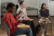© pressmaster - Three women sitting in chairs attending group therapy session, Black young adult woman reading brochure, Caucasian middle aged woman and Hispanic woman listening attentively