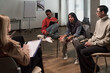 © pressmaster - Diverse group of young adults and middle aged women sitting in circle discussing during group therapy session, multiethnic participants engaging in conversation indoors