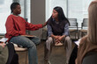 © pressmaster - Young Black woman comforting Hispanic woman during group therapy session, both sitting on chairs, other participants visible in foreground, emotional support interaction