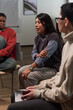 © pressmaster - Multiethnic group of adults sitting in circle participating in group discussion, Hispanic woman speaking while Black woman and Caucasian woman listening attentively in modern room
