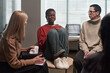 © pressmaster - Black woman sitting with knees to chest appearing emotional during group therapy session with Caucasian woman holding tissue box, and mature Caucasian woman listening nearby