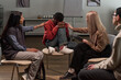 © pressmaster - Black teenager sitting curled up on chair, covering face with hands while Caucasian woman and Hispanic woman sitting nearby offering comfort and support during group therapy session
