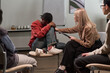 © pressmaster - Black woman sitting with head down on knees while Caucasian woman offering comfort by touching arm during group therapy session, with other people present in background