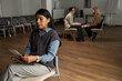 © pressmaster - Hispanic woman sitting alone reading leaflet in foreground while two Caucasian women sitting together having conversation in background in empty meeting room, group therapy session concept