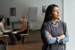 © pressmaster - Hispanic woman standing with arms crossed looking out window, two Caucasian adults sitting in background having conversation in modern psychologists office setting