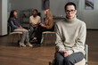 © pressmaster - Portrait of middle aged Caucasian woman with short dark hair wearing glasses sitting in foreground looking at camera, diverse group of adults talking in background during support meeting