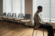 © pressmaster - Depressed mature Caucasian woman sitting alone on chair near window with blinds, looking down with hands clasped, empty chairs lined up in background, wooden floor visible