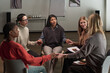 © pressmaster - Diverse group of middle aged and young adult women sitting in circle holding hands, supporting each other during group therapy session in modern room with wooden floor