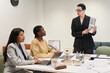 © pressmaster - Middle aged Caucasian woman standing and presenting document to young Black woman and young Hispanic woman sitting at conference table, discussing business strategy during meeting