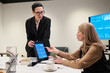 © pressmaster - Caucasian middle aged woman sitting at table discussing financial data on digital tablet with standing colleague in business suit during office meeting