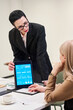 © pressmaster - Middle aged woman with short dark hair and glasses standing and presenting digital tablet with analytics dashboard to seated Caucasian woman during business meeting