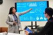 © pressmaster - Young adult Hispanic woman presenting data on digital screen to Caucasian woman during business meeting, gesturing toward charts while colleague taking notes on tablet