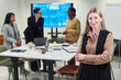 © pressmaster - Portrait of Caucasian middle aged woman standing with arms crossed in foreground, diverse group of colleagues discussing business strategy near digital screen in office