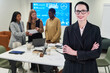 © pressmaster - Portrait of middle aged Caucasian woman smiling with arms crossed in foreground, diverse group of women collaborating on business project with digital screen in background