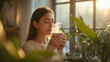 © siraphol - Young woman enjoying warm drink in sunlit plant filled room, serene morning comfort
