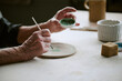 © AnnaStills - Senior Caucasian woman painting ceramic plate with brush, holding small bowl of green paint, showing detailed hand movements and creative process at table in studio