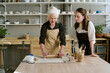 © AnnaStills - Senior Caucasian woman demonstrating pottery technique to young adult Caucasian woman in ceramics studio, both standing at worktable with clay tools and pottery materials visible