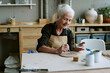 © AnnaStills - Senior Caucasian woman painting ceramic plate at table, holding paintbrush and small container, smiling while working on pottery project in creative studio environment