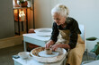 © AnnaStills - Senior Caucasian woman shaping clay on pottery wheel, concentrating on creating ceramic vessel with hands, sitting at workstation in studio, demonstrating pottery making process