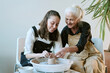 © AnnaStills - Caucasian young adult woman and Caucasian senior woman shaping clay together on pottery wheel, both smiling and focusing on hands while creating ceramic piece in studio setting
