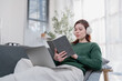 © Tj - Relaxed woman wearing headphones sits comfortably on a couch reading a book beside a laptop, combining study and leisure in a bright modern home interior