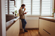© Dragana Gordic - Casual Man Drinking Coffee and Checking Smartphone While Standing in Bright Home Kitchen