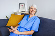 © Xavier Lorenzo - Senior healthcare worker relaxing on sofa using mobile phone after workday. Social media and healthcare concept