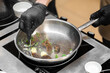 © pavel siamionov - A professional chef in black gloves sautés gourmet morel mushrooms with fresh butter and herbs in a stainless steel pan on a modern induction stovetop in a commercial kitchen.