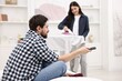 © New Africa - Gender inequality concept. Man with remote control sitting on bed while his wife ironing clothes indoors, selective focus