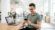 © ryker - Smiling man in sage green polo shirt using smartphone at modern bright office desk with laptop and coffee, casual professional scene with natural light and plants