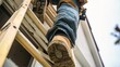 © ds17 - A close-up view of a worker's feet on a wooden ladder. The worker wears blue jeans and brown work boots. The background shows a house under maintenance.