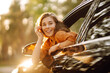 © maxbelchenko - A smiling woman leans out of a car window and enjoys the sunset. A beautiful woman on a car adventure feels free in the sun's rays. The concept of freedom, adventure, and the car.